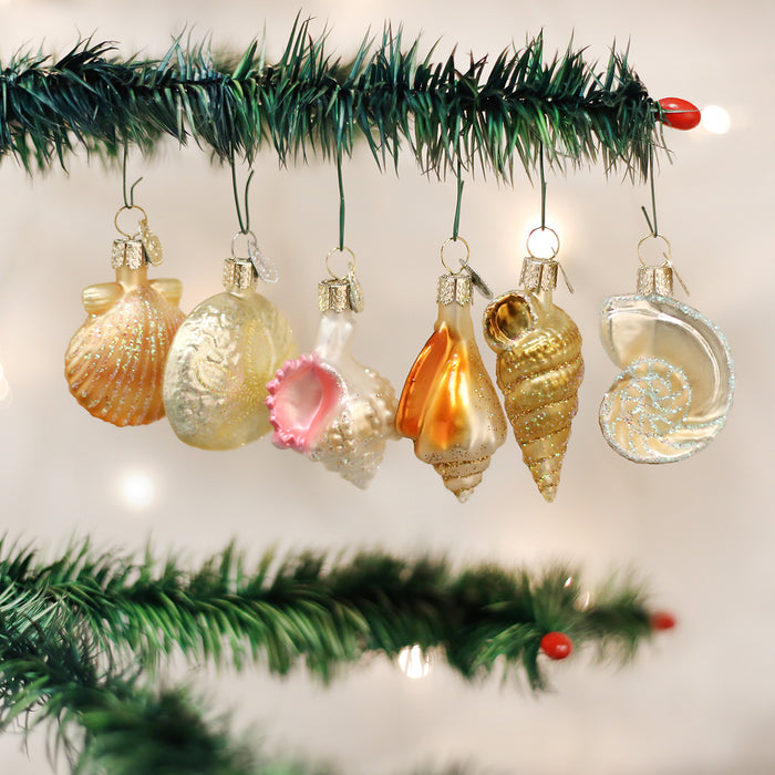 Seven glass Christmas ornaments shaped like seashells hang on a green pine branch, with soft white holiday lights blurred in the background.