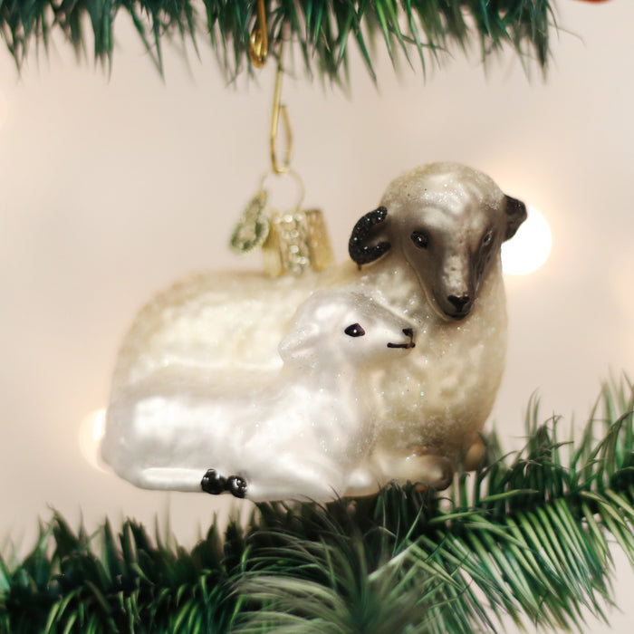 A Christmas tree ornament shaped like a brown and white sheep with a lamb resting beside it, hanging on a green tree branch with blurred lights in the background.