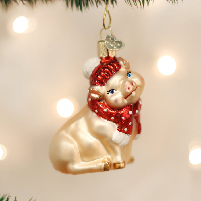 A shiny pig ornament wearing a red scarf and knit hat hangs from a Christmas tree branch, with soft white holiday lights glowing in the blurred background.