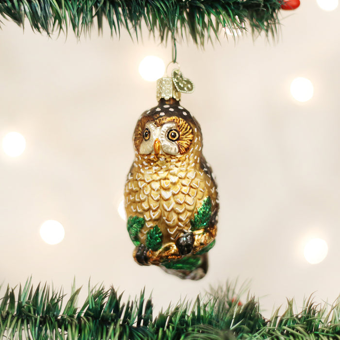 A shiny owl-shaped Christmas ornament hangs from a green pine branch, with blurred white lights in the background.