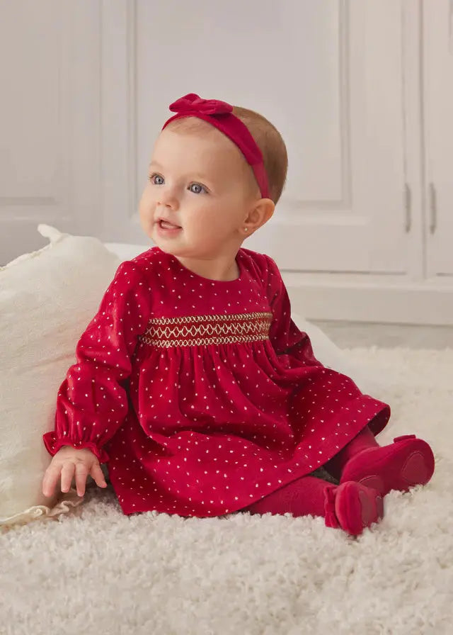 Seated baby girl wearing red Mayoral velvet dress with smocked bodice