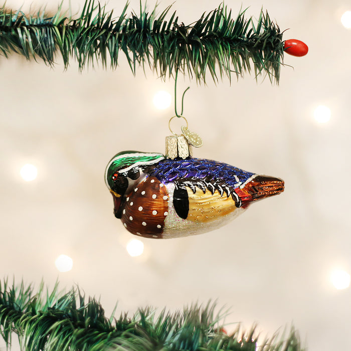 A colorful bird-shaped Christmas ornament hangs from a green tree branch, with soft white lights glowing in the blurred background.